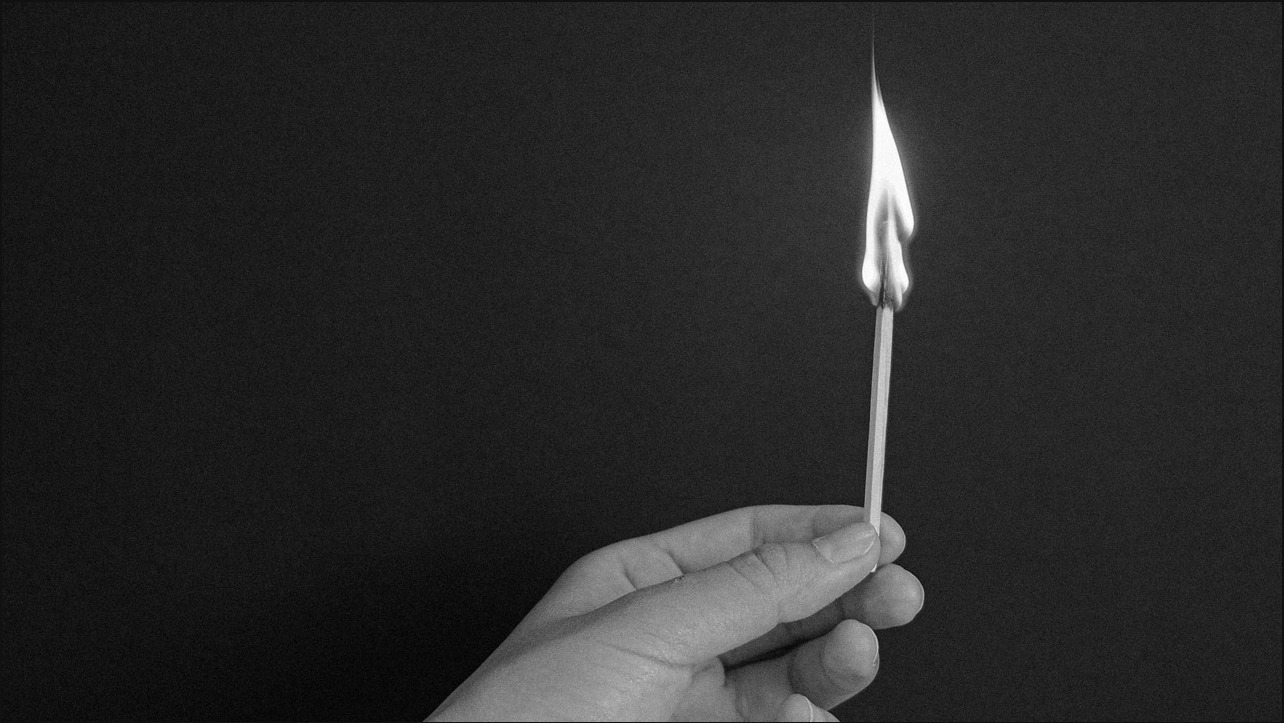A hand holding a single burning match against darkness